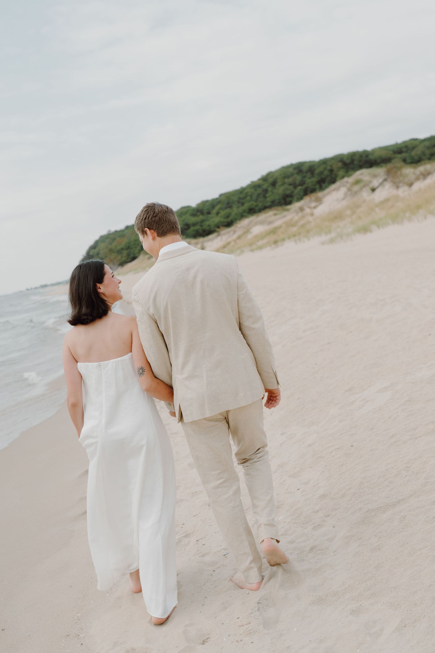 A couple in matching light colored outfits walks together on a peaceful beach.