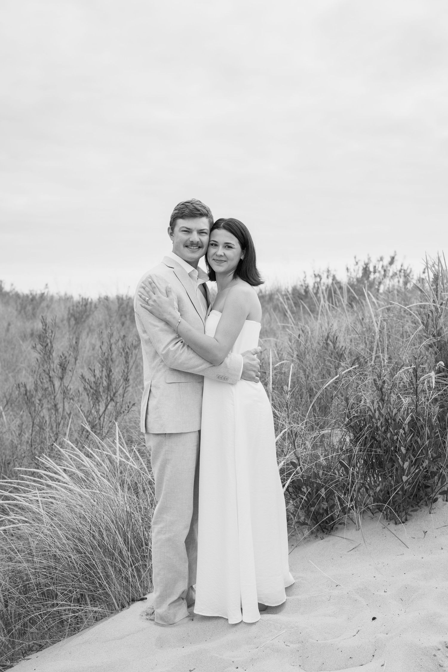 Black and white wedding portraits showing joyful couple embracing on coastal dunes.