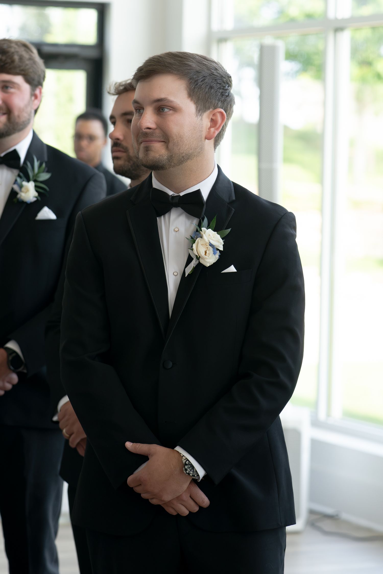 Groom in black tuxedo with white boutonniere stands at altar, groomsmen visible behind him during wedding ceremony.