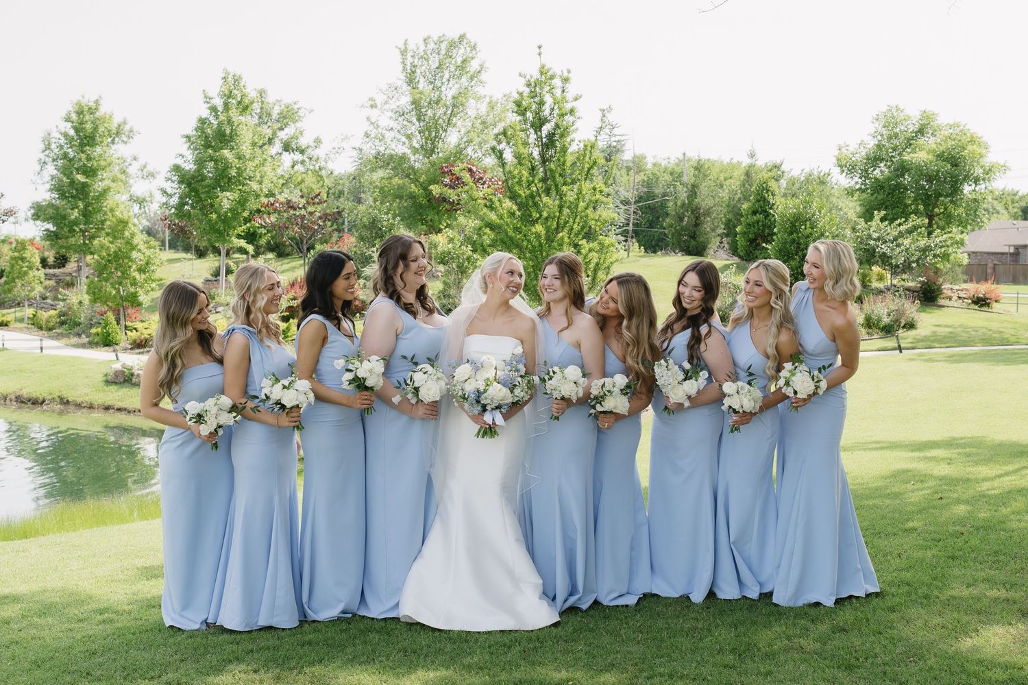 Bride in white gown poses with ten bridesmaids in light blue dresses holding white bouquets on a green lawn.