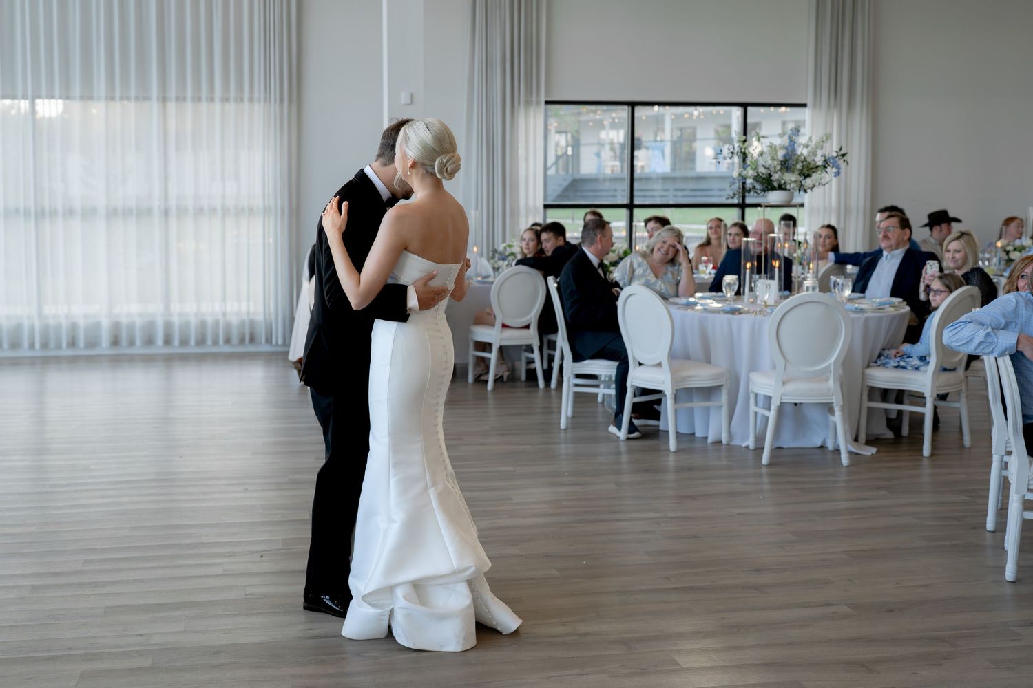Bride and groom share first dance in elegant white wedding reception hall with guests seated at round tables.