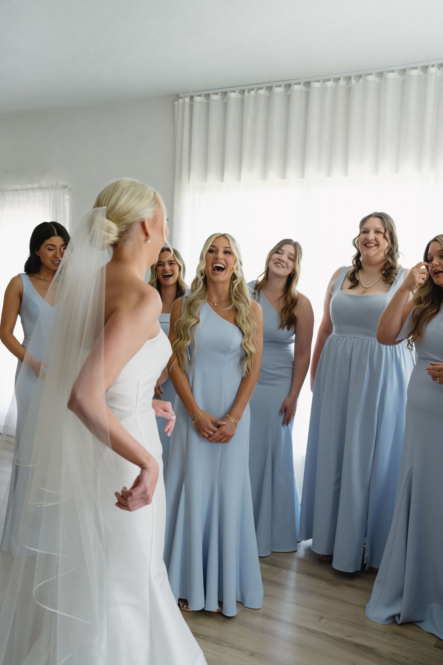 Bride in white gown and veil reveals look to bridesmaids in light blue dresses, who react with joy and excitement.