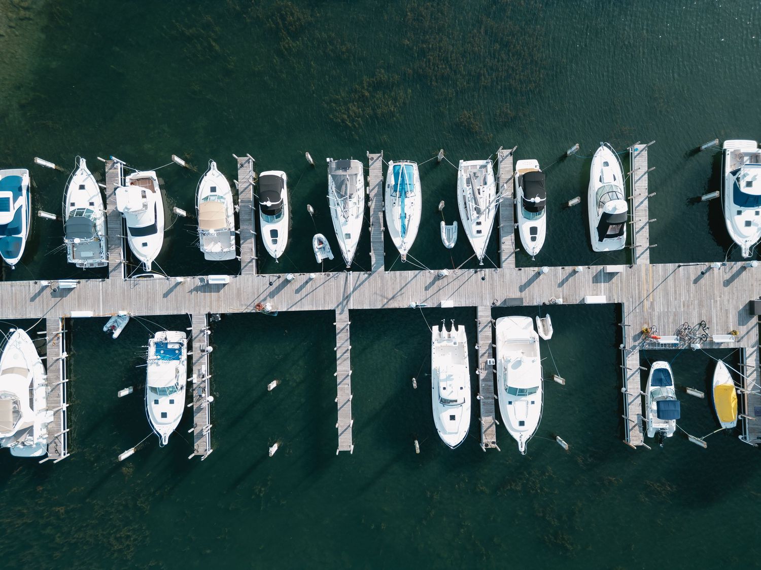 Aerial view of multiple white boats docked at a marina pier surrounded by dark green water.