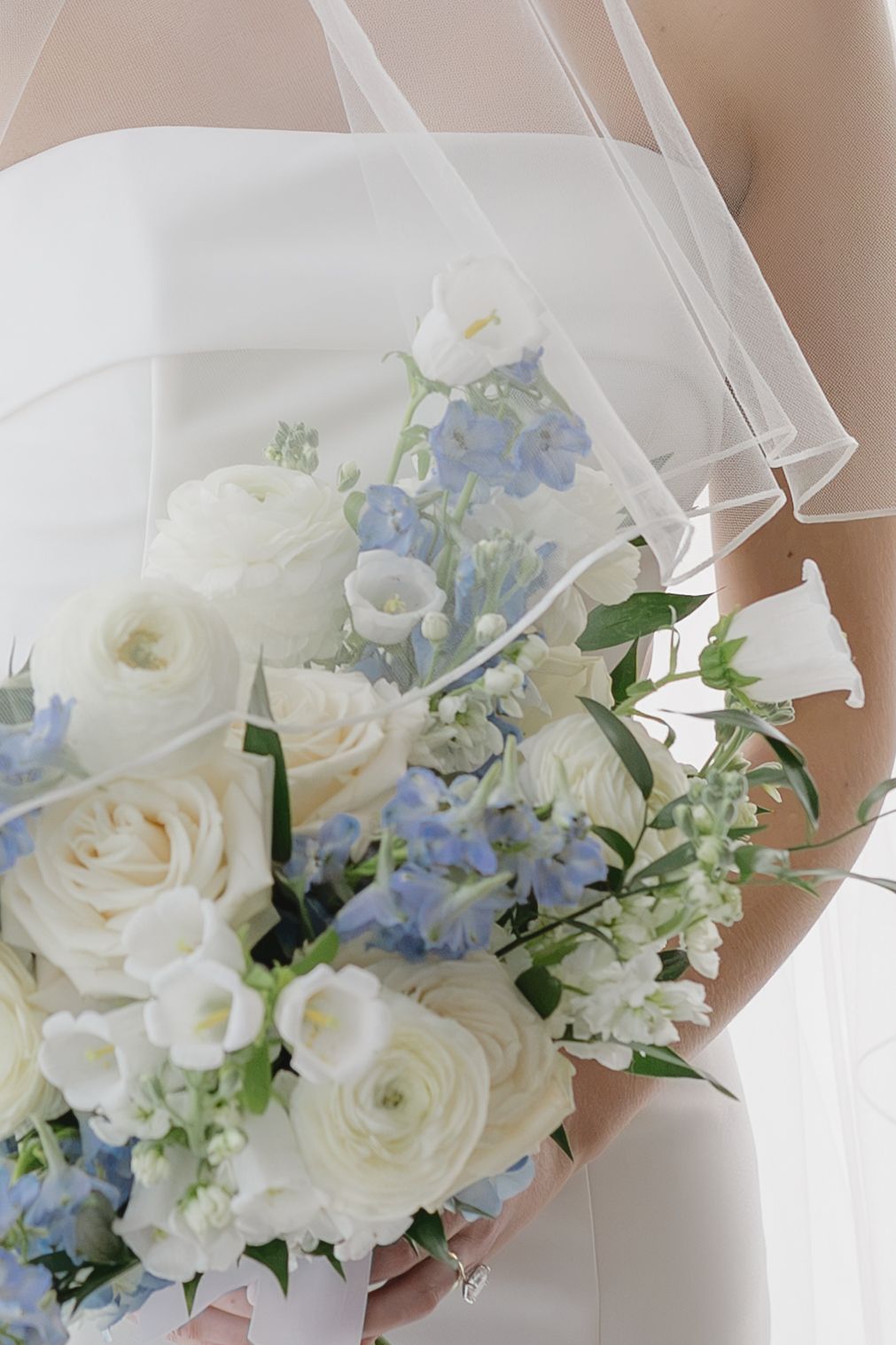 Bride holding elegant wedding bouquet of white roses, ranunculus, and blue delphinium flowers under a sheer veil.
