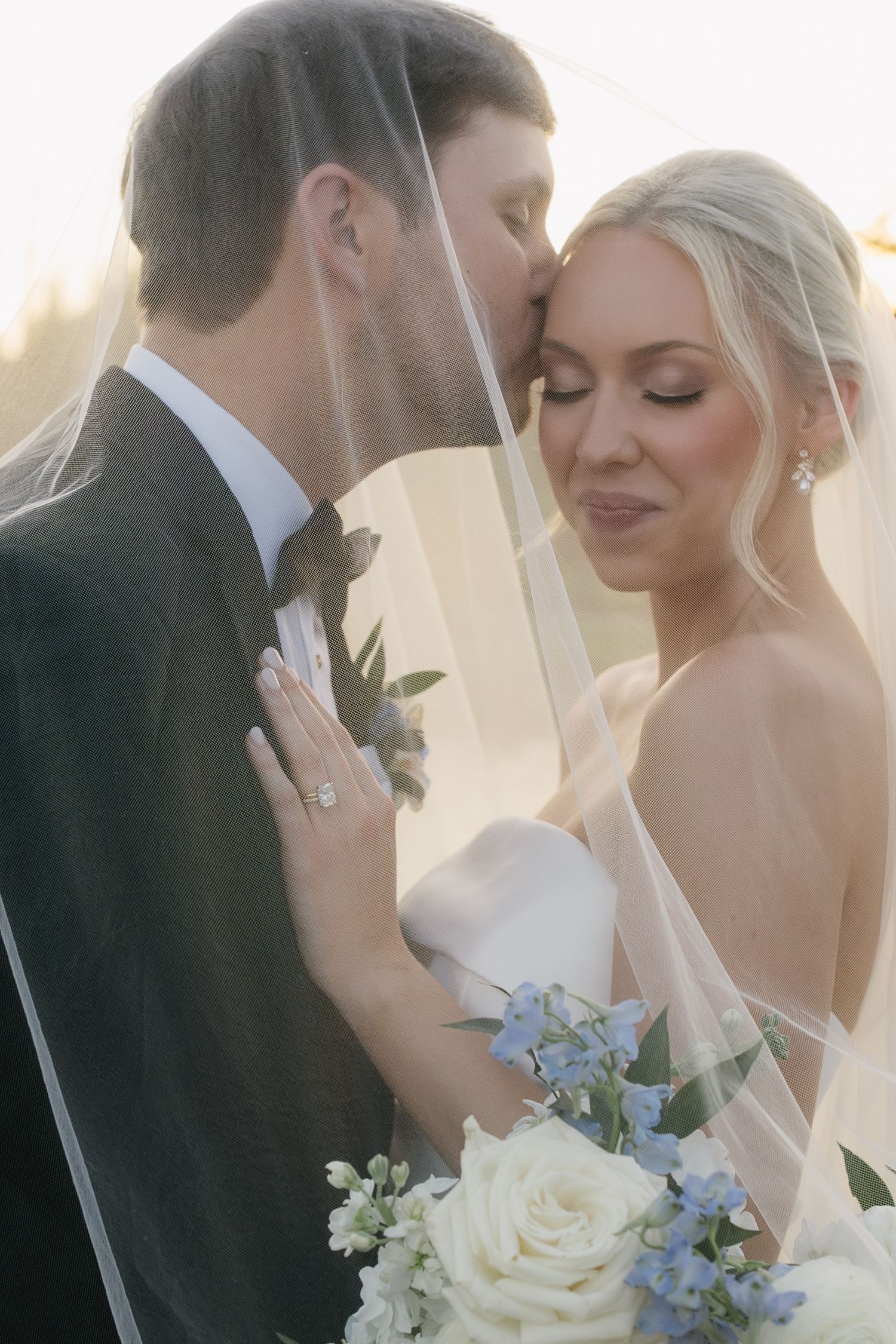 Groom kissing bride on forehead under veil, bride holding white and blue bouquet in romantic golden hour wedding portrait.