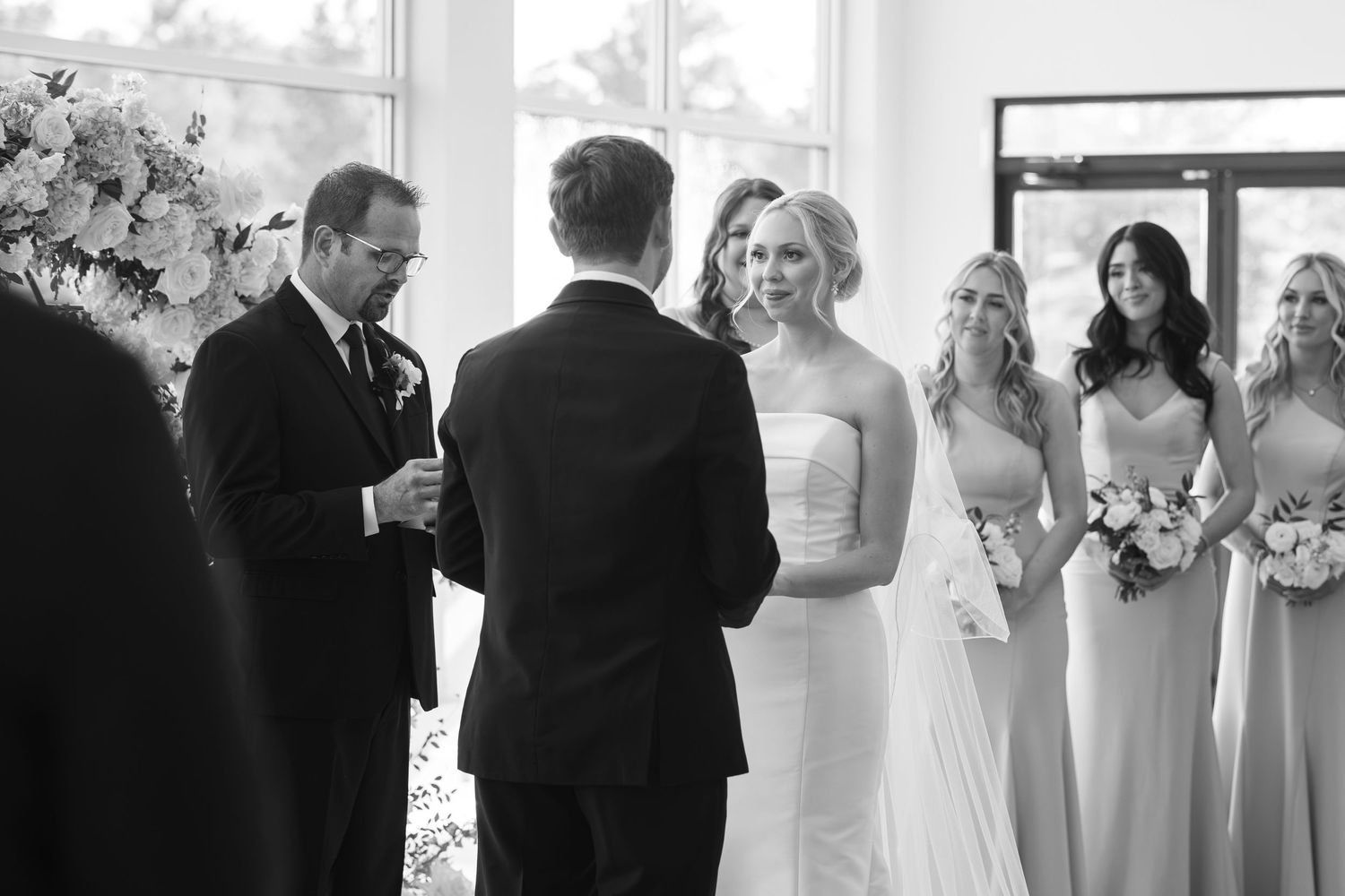 Black and white wedding ceremony photo with bride, groom, officiant, and bridesmaids holding bouquets.
