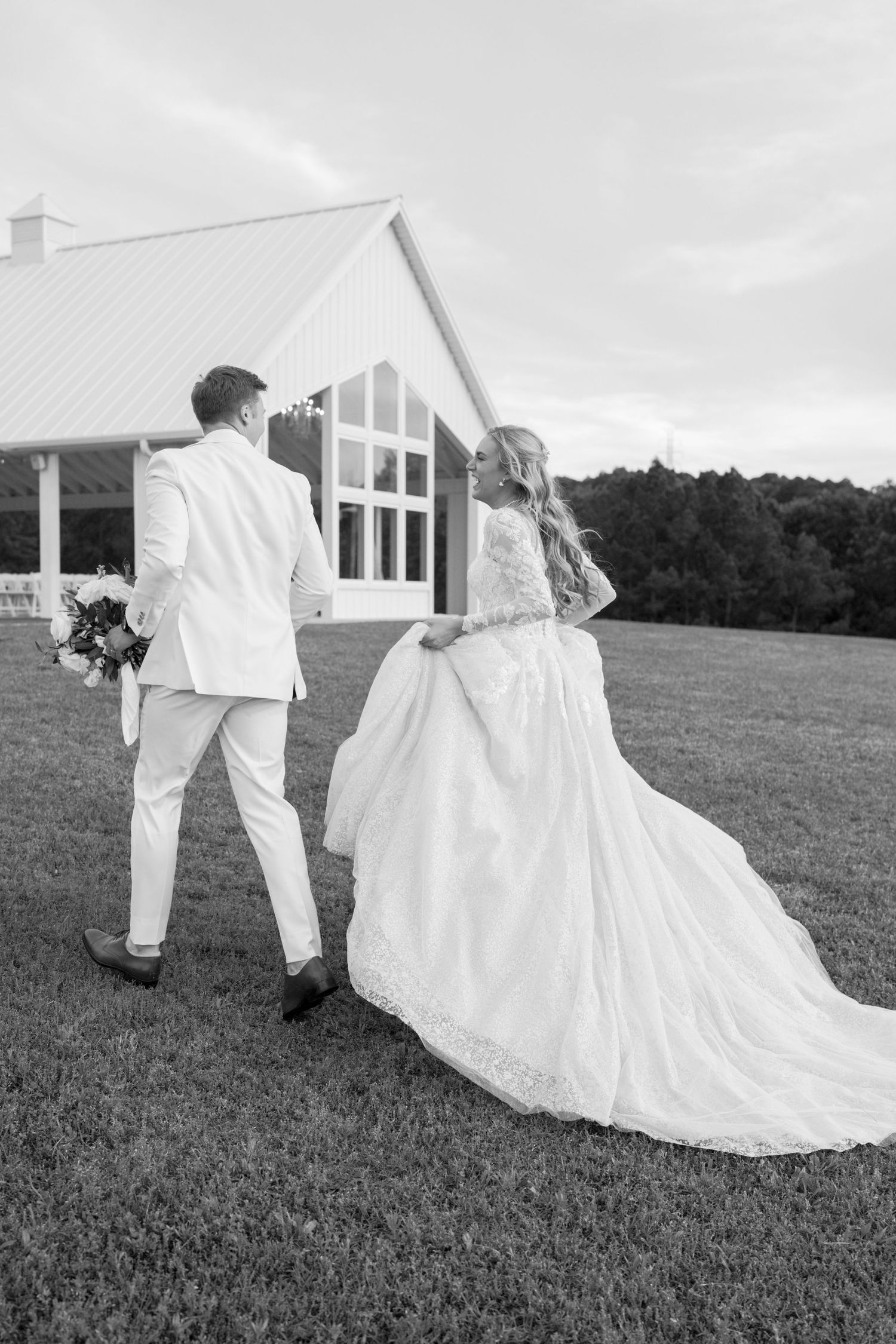 A bride in a flowing white gown and groom in white suit walk across a grassy lawn towards a modern white barn venue.