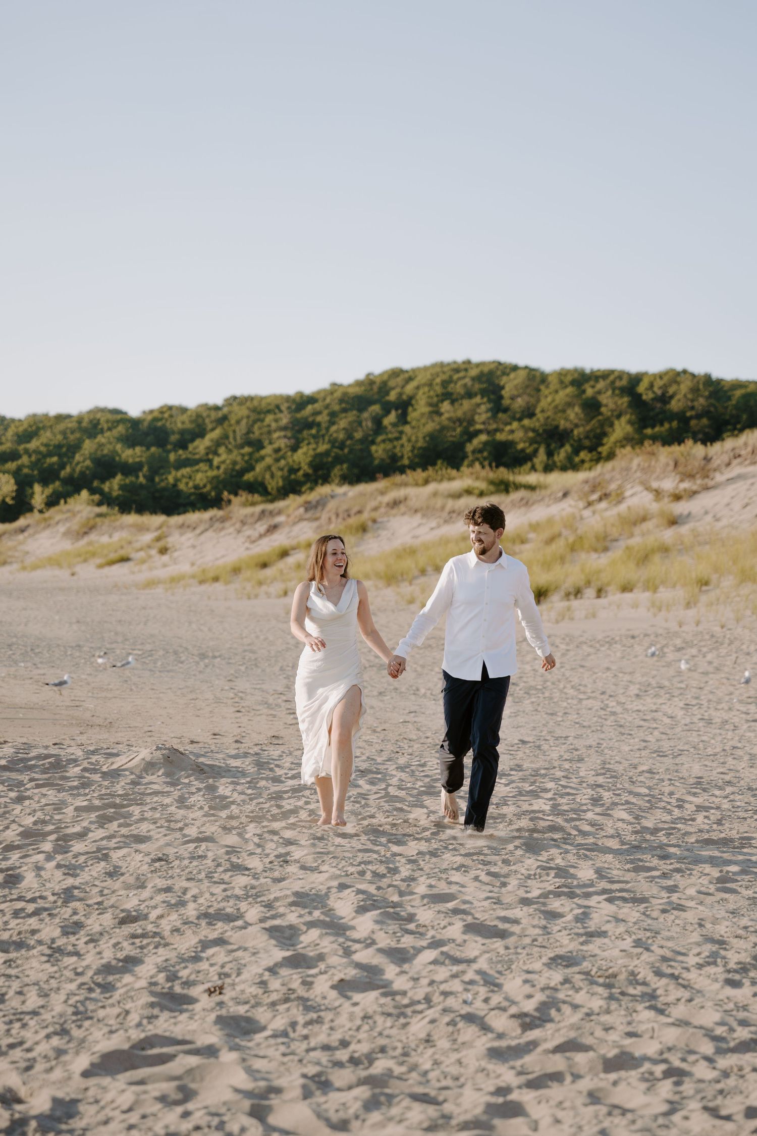 A couple runs playfully together on a sandy beach with grassy dunes in the background.