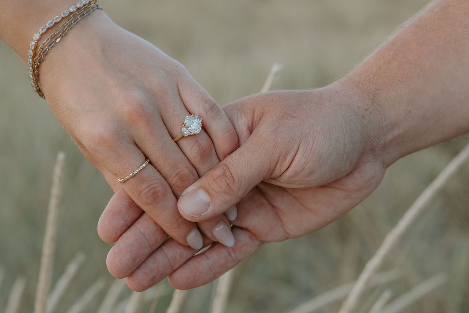 Close-up of hands with engagement rings touching against blurred beach grass background.