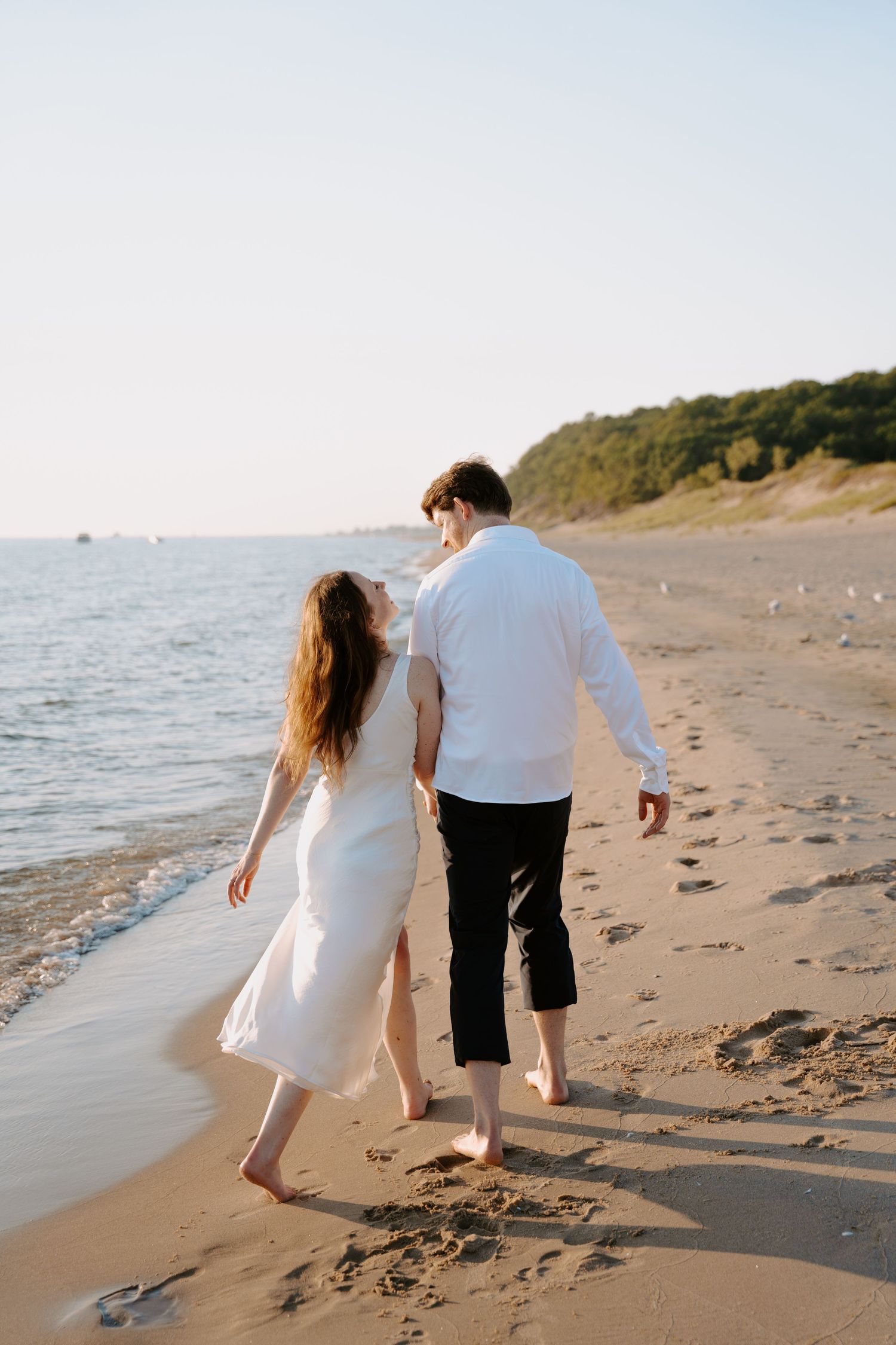 Couple walking barefoot on a sandy beach with gentle waves beside them.