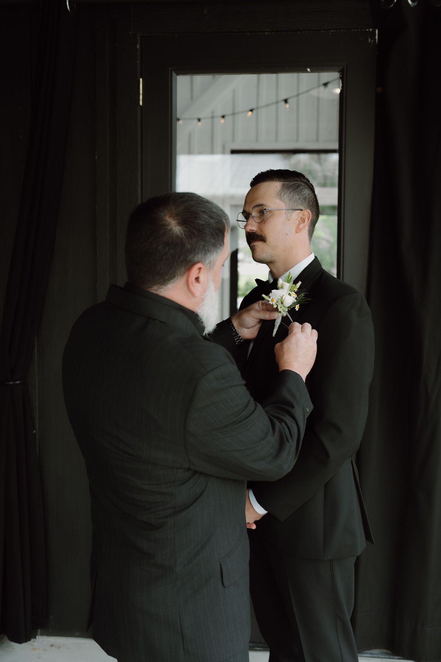 Groomsmen help adjust boutonnieres and ties before a formal event in a dark interior setting.