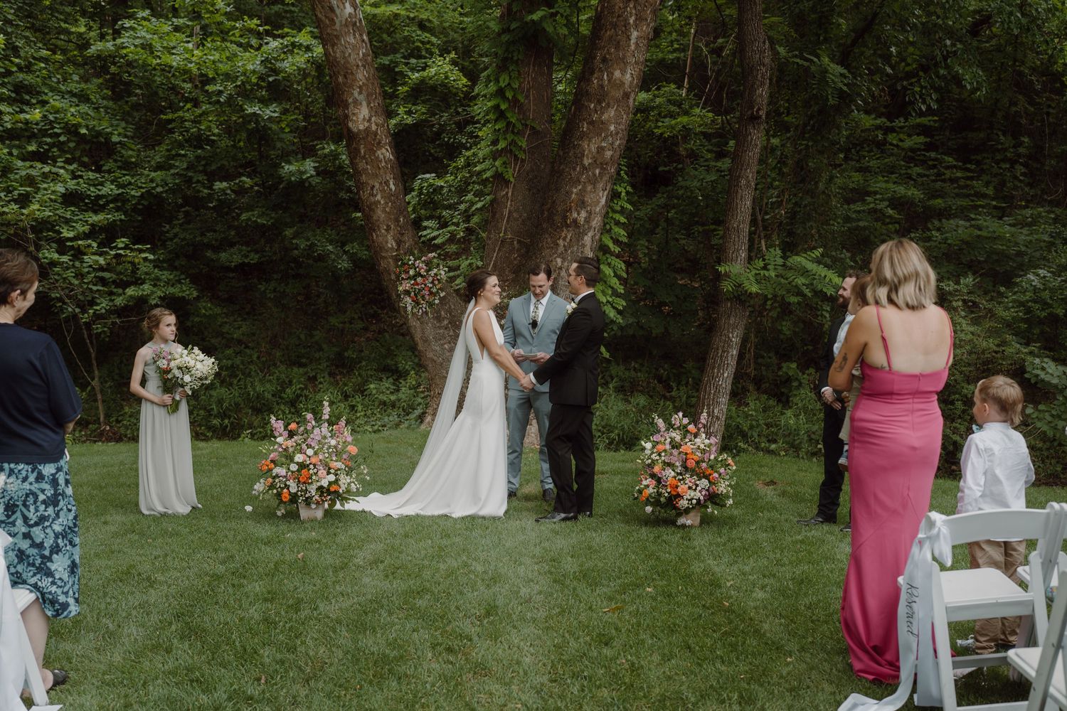 Wedding guests and party members mingle in a scenic wooded outdoor venue.