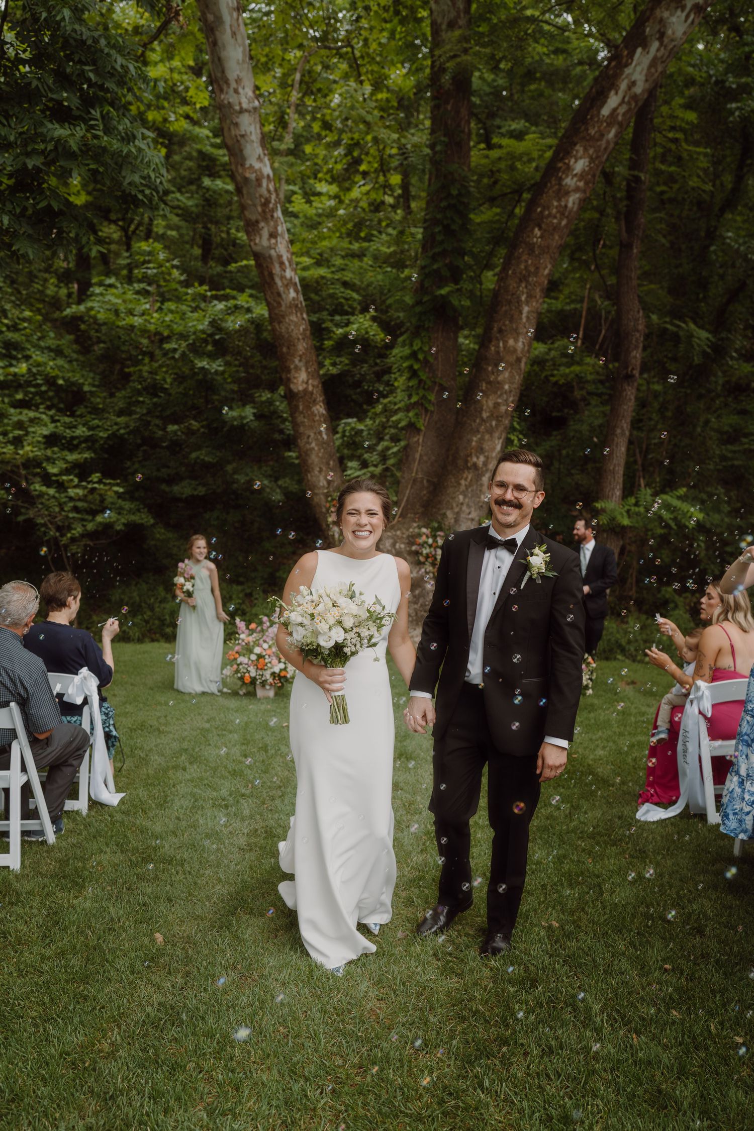 Wedding party processes down a grassy aisle between white chairs in a woodland setting.