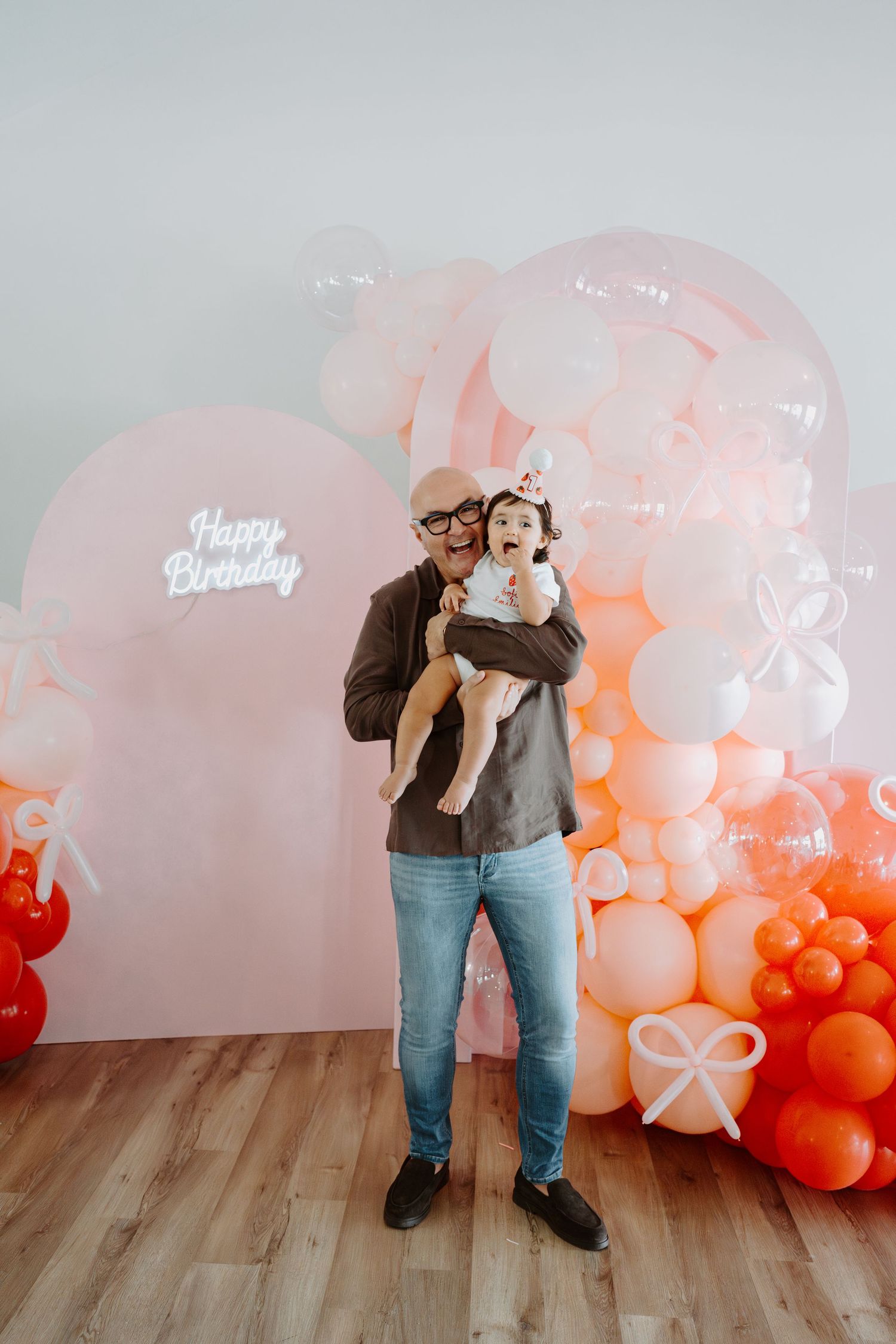 Festive birthday setup with ombre coral balloon arch and 'Happy Birthday' sign on white wall.