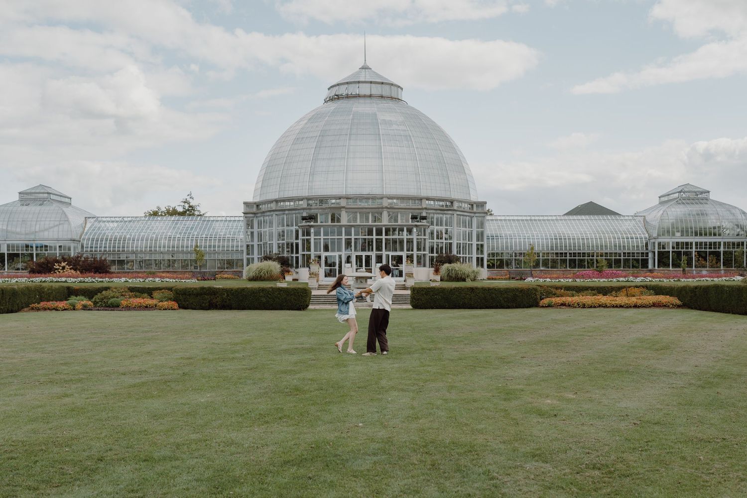 Two people standing on green lawn with symmetrical glass conservatory building behind them.