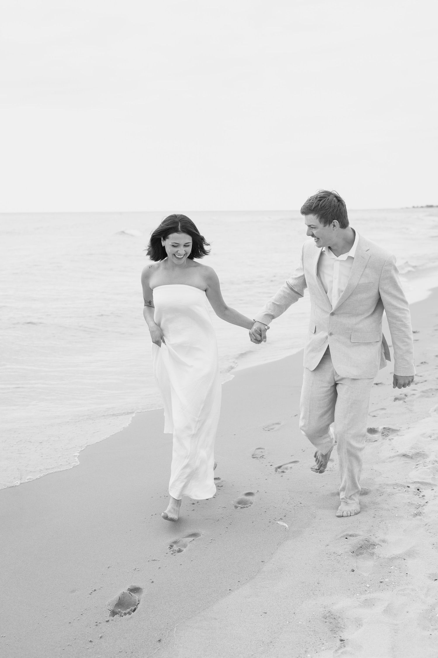 Two people in light colored outfits run hand in hand along a peaceful beach shore.