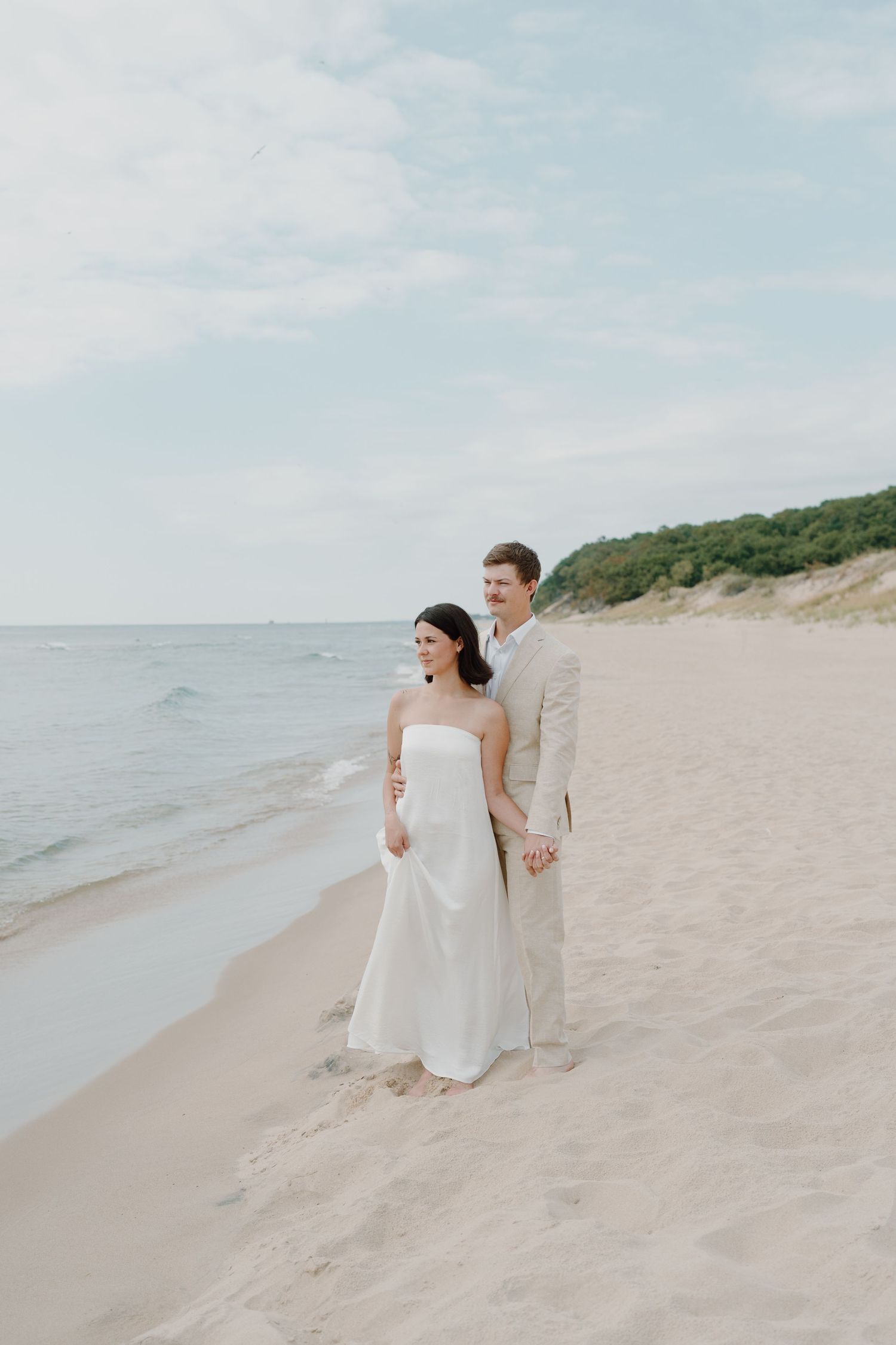 A couple in wedding attire stands together on a peaceful beach with rolling dunes in the background.