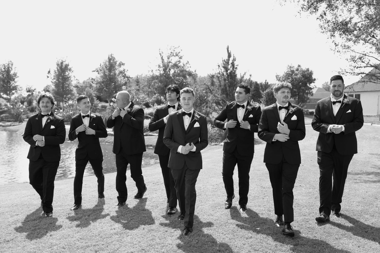 Groomsmen in black tuxedos strike playful jumping poses outdoors for wedding photos.