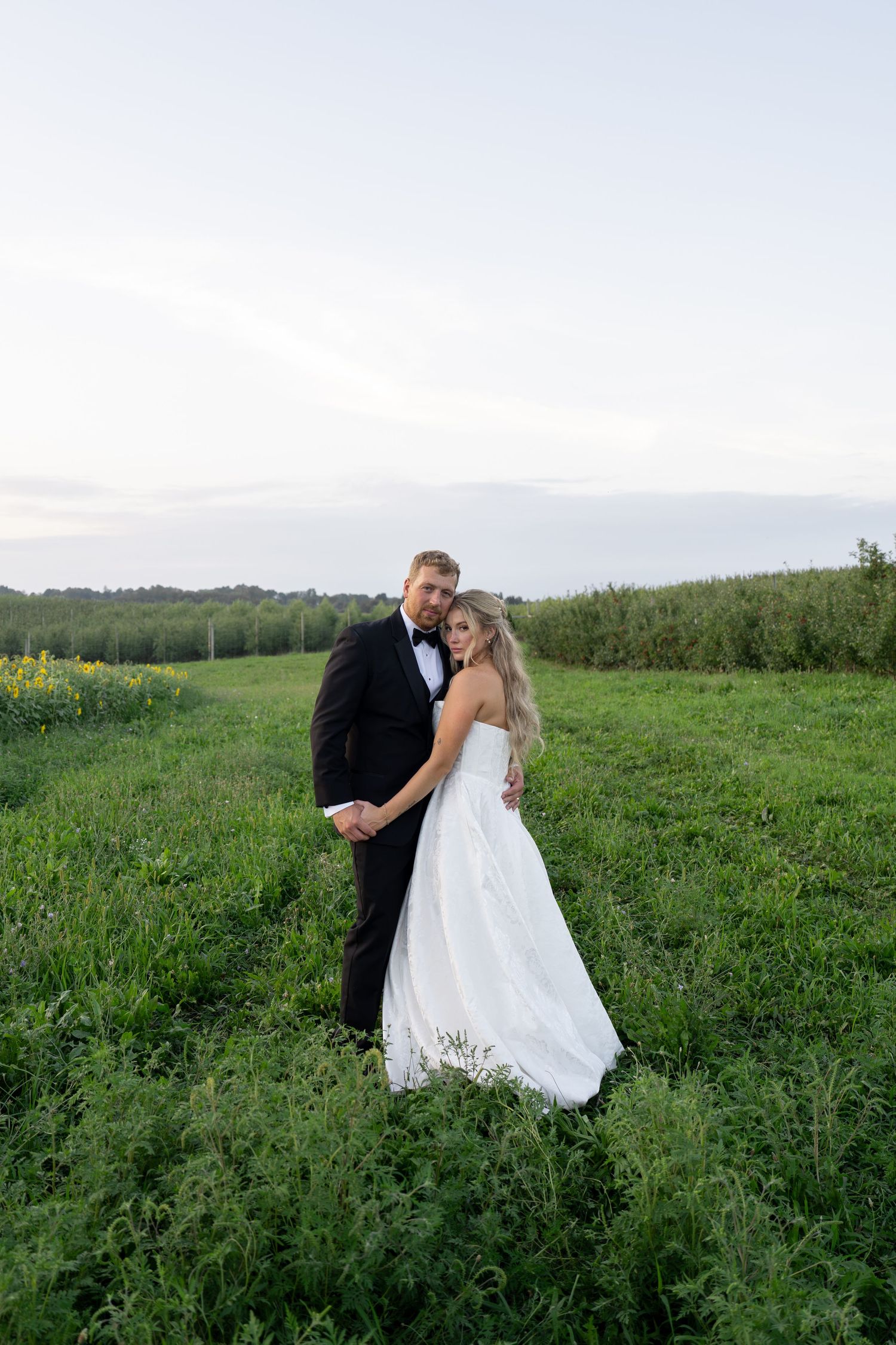 A bride and groom embrace in a lush green field at dusk, wearing a white gown and black tuxedo.