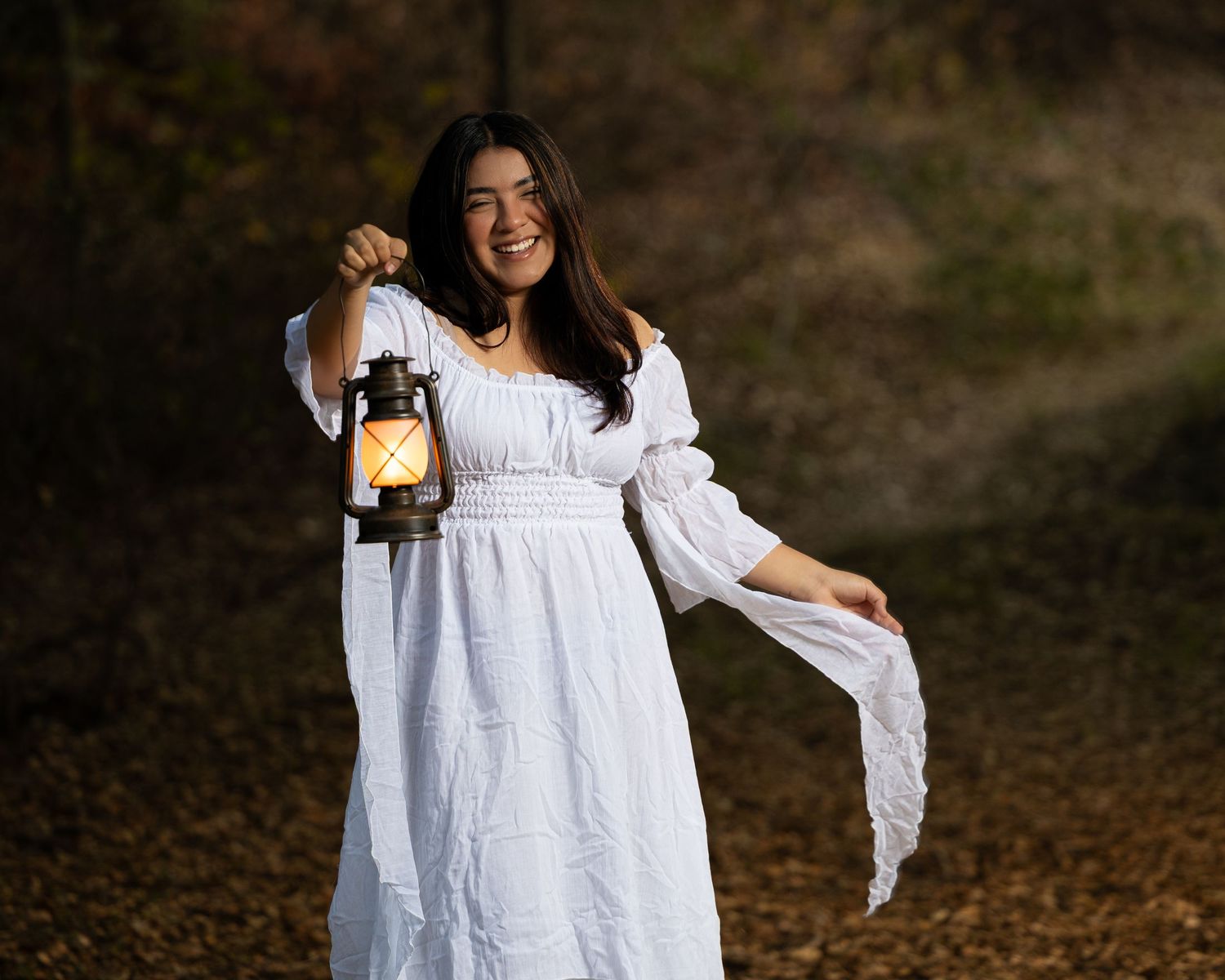 A woman in a flowing white dress holds a vintage lantern while walking through autumn leaves in a forest.