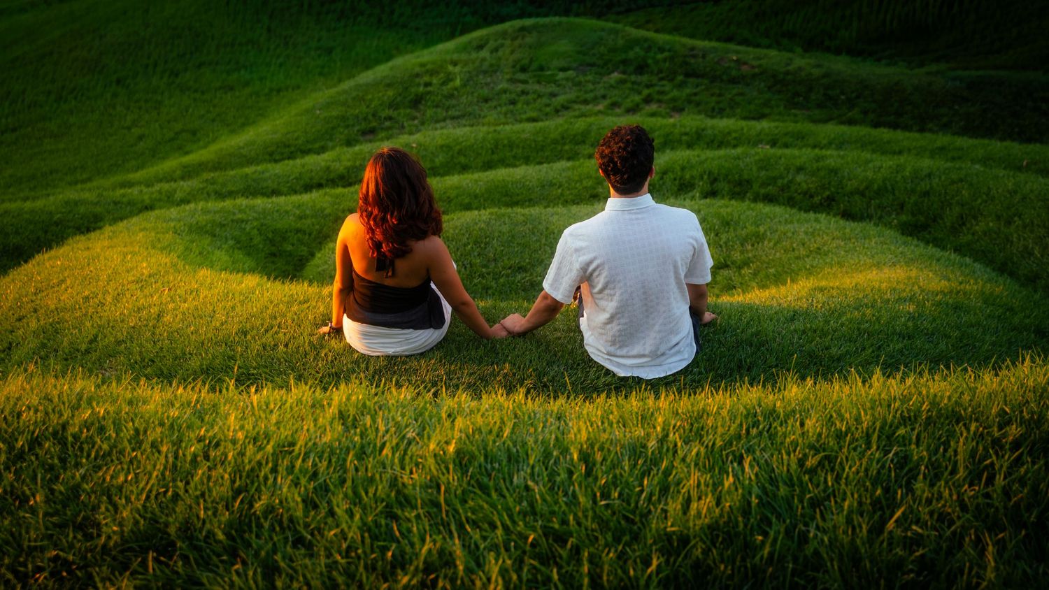 A couple sits hand in hand on lush green rolling hills, enjoying a serene moment together in nature.