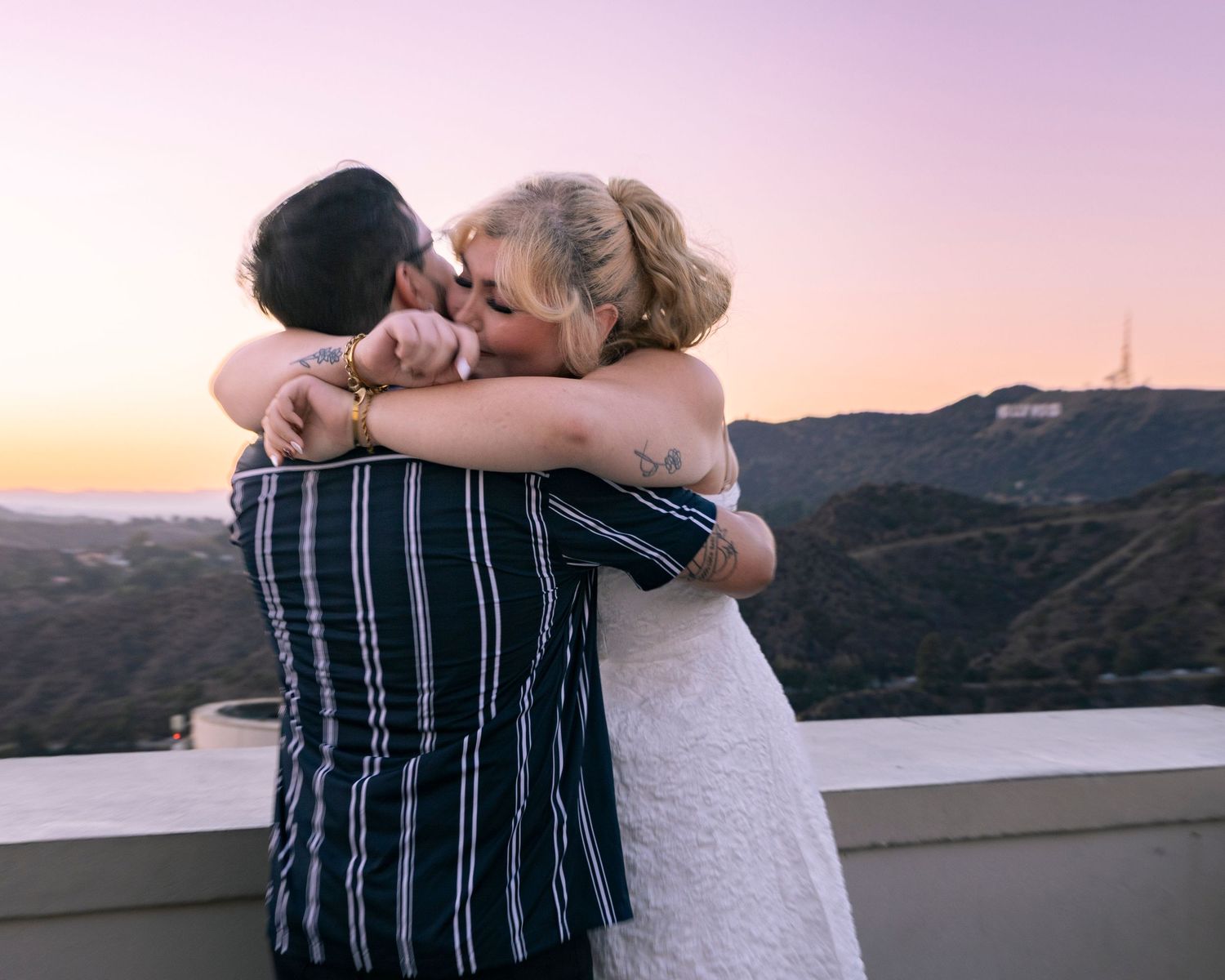 A couple shares an intimate moment against a sunset backdrop overlooking mountains and water.