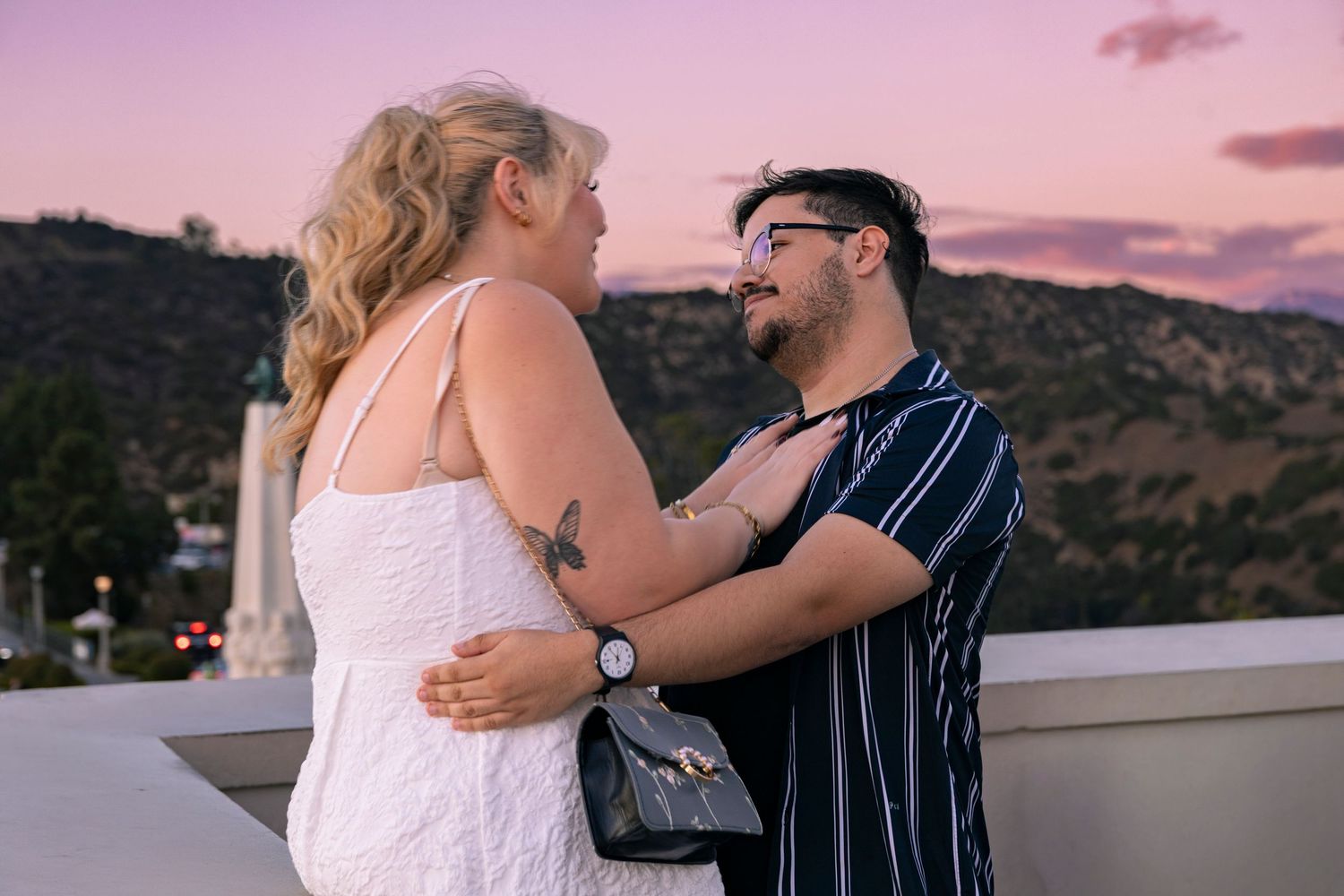 A couple shares a romantic embrace on a balcony during sunset with mountains in the background.