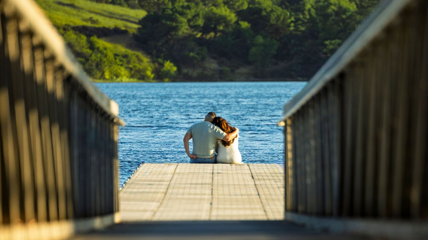 Wooden dock walkway leads to a sunny lakeside pier with blue water and green trees in the background.
