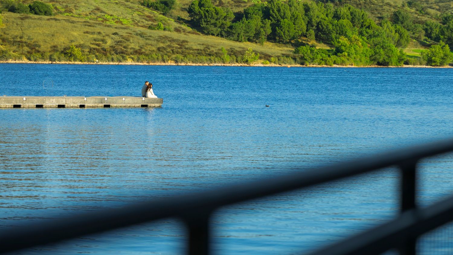 A panoramic view of a calm blue lake surrounded by lush green hills with a stone pier extending into the water.