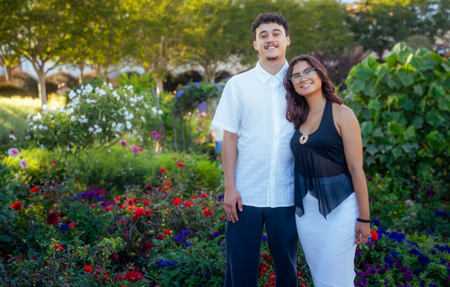 A smiling couple poses together in a vibrant garden full of colorful flowers on a sunny day.