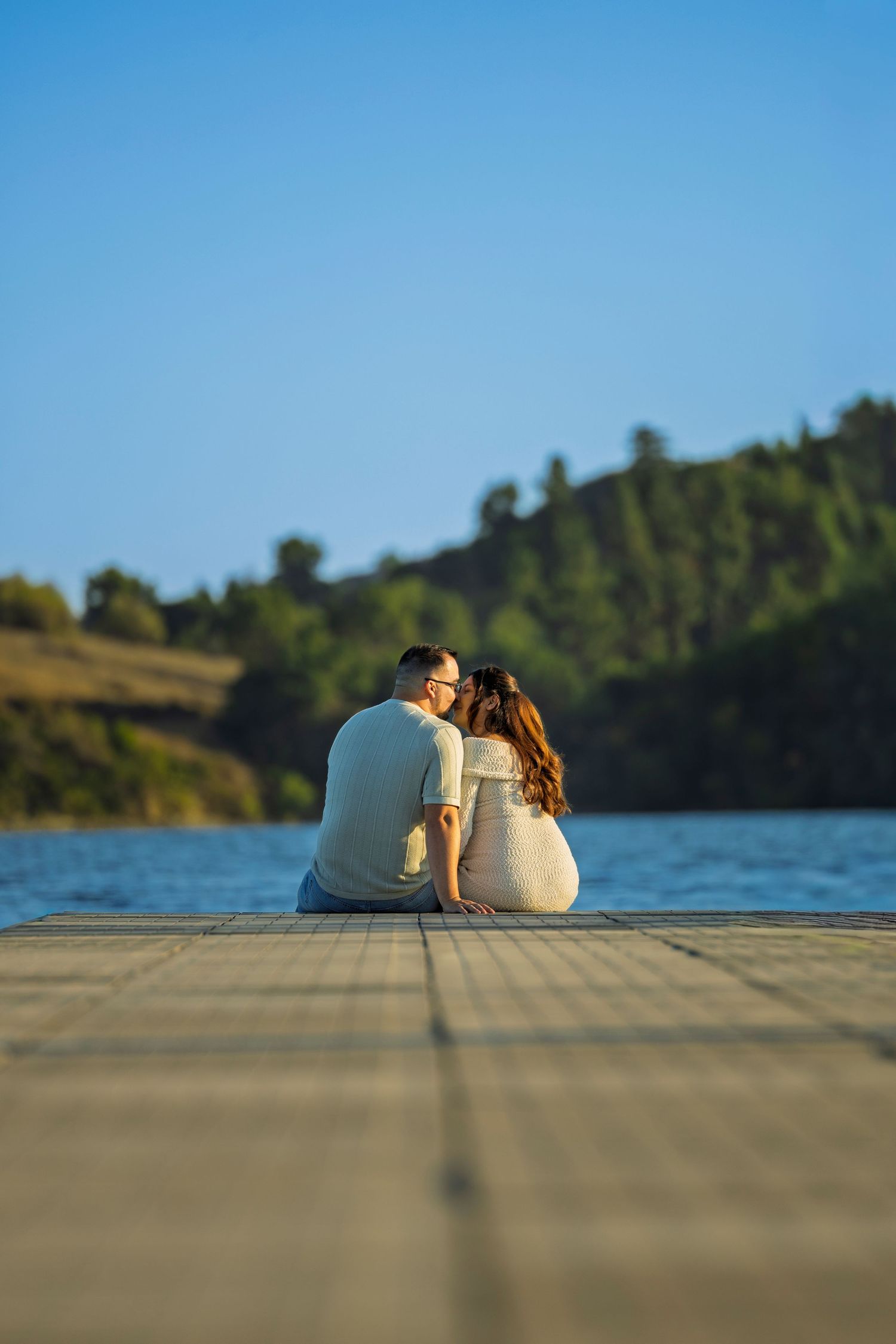 A couple sits close together on a wooden dock by a serene lake surrounded by trees on a sunny day.