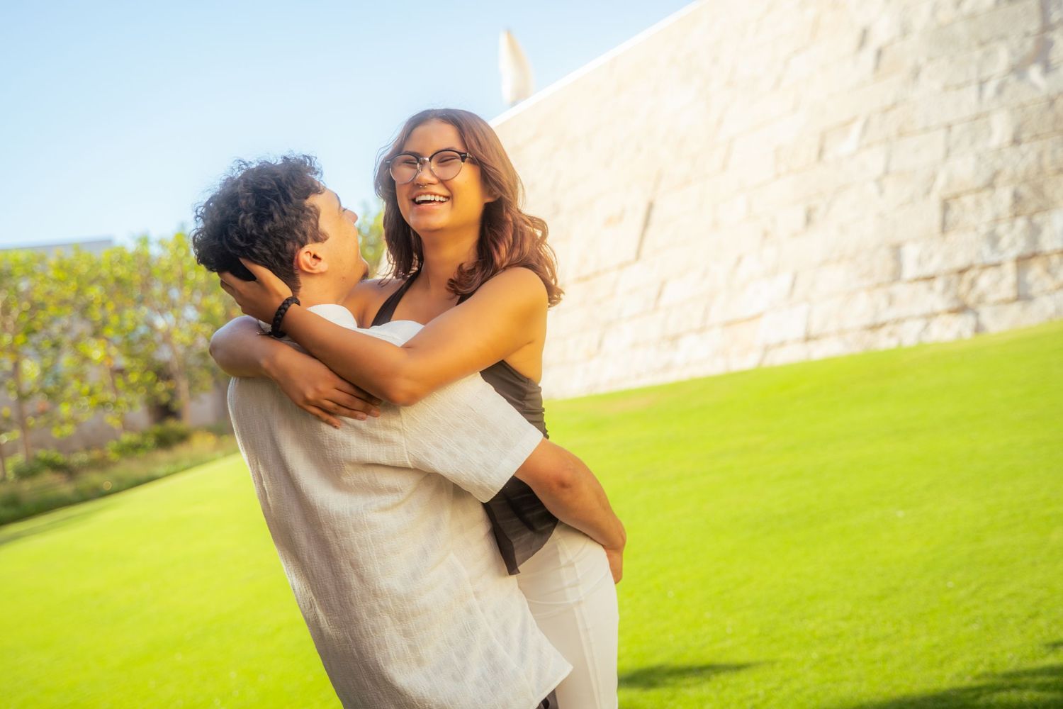 Happy couple embracing and laughing outdoors on green grass near a stone wall on a sunny day.