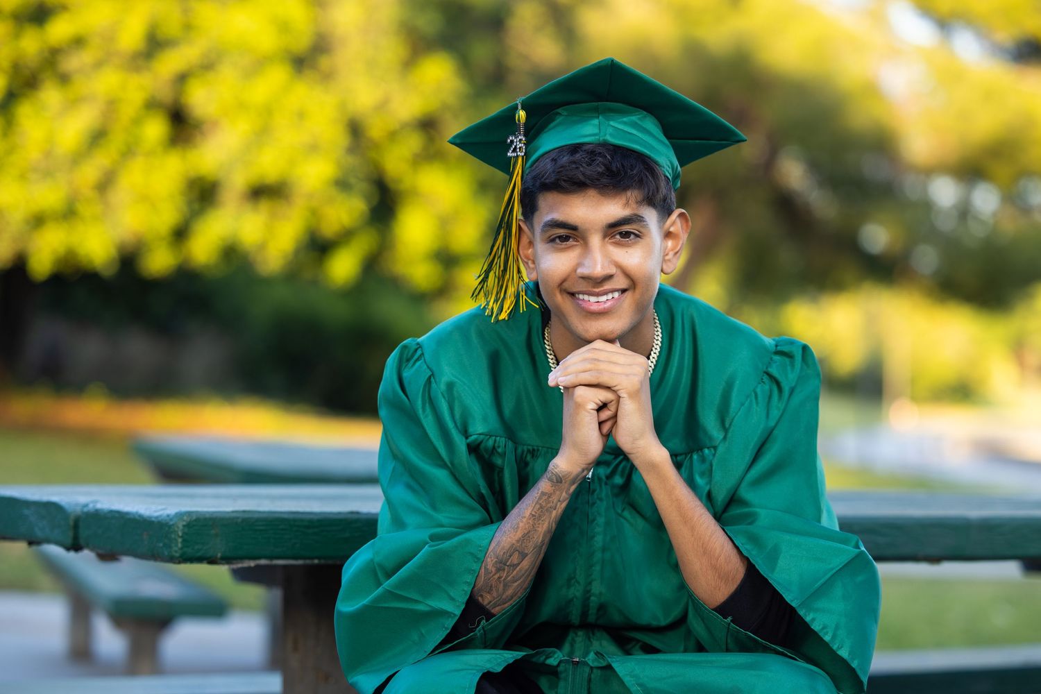 Smiling young man in green graduation cap and gown sitting outdoors at a picnic table with trees in background.