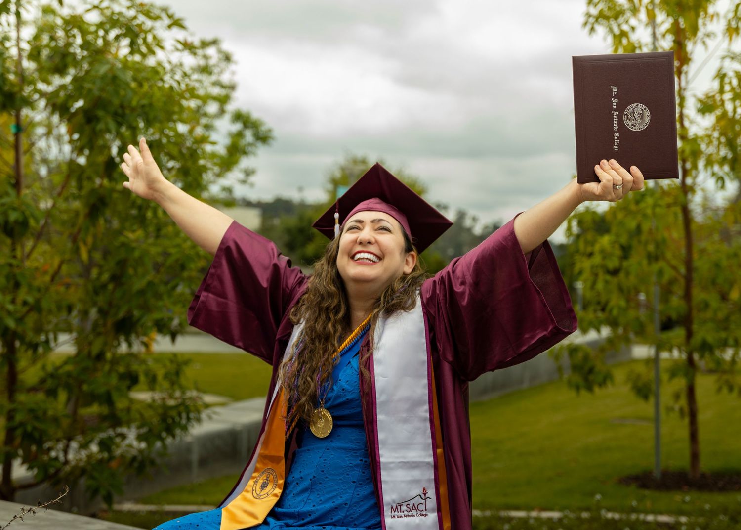 Graduate celebrates achievement outdoors on a cloudy day with trees in background.