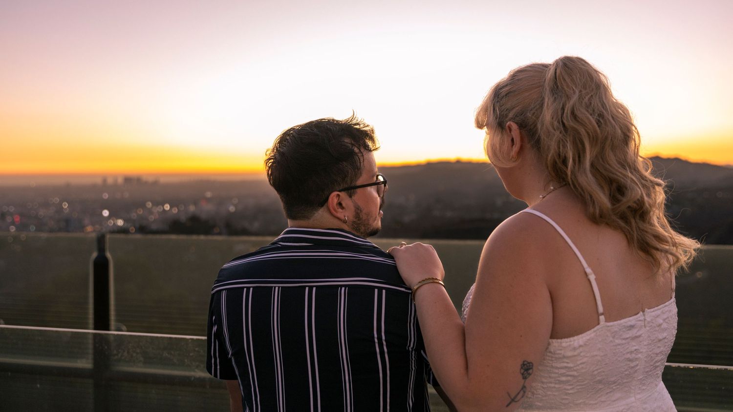 A couple shares a romantic moment at sunset overlooking a cityscape from a scenic viewpoint.