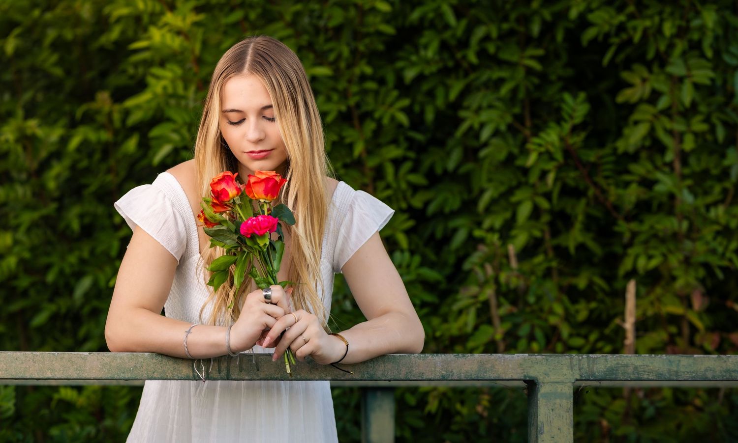 Young blonde woman in white dress holding red roses bouquet, leaning on railing with green foliage background.