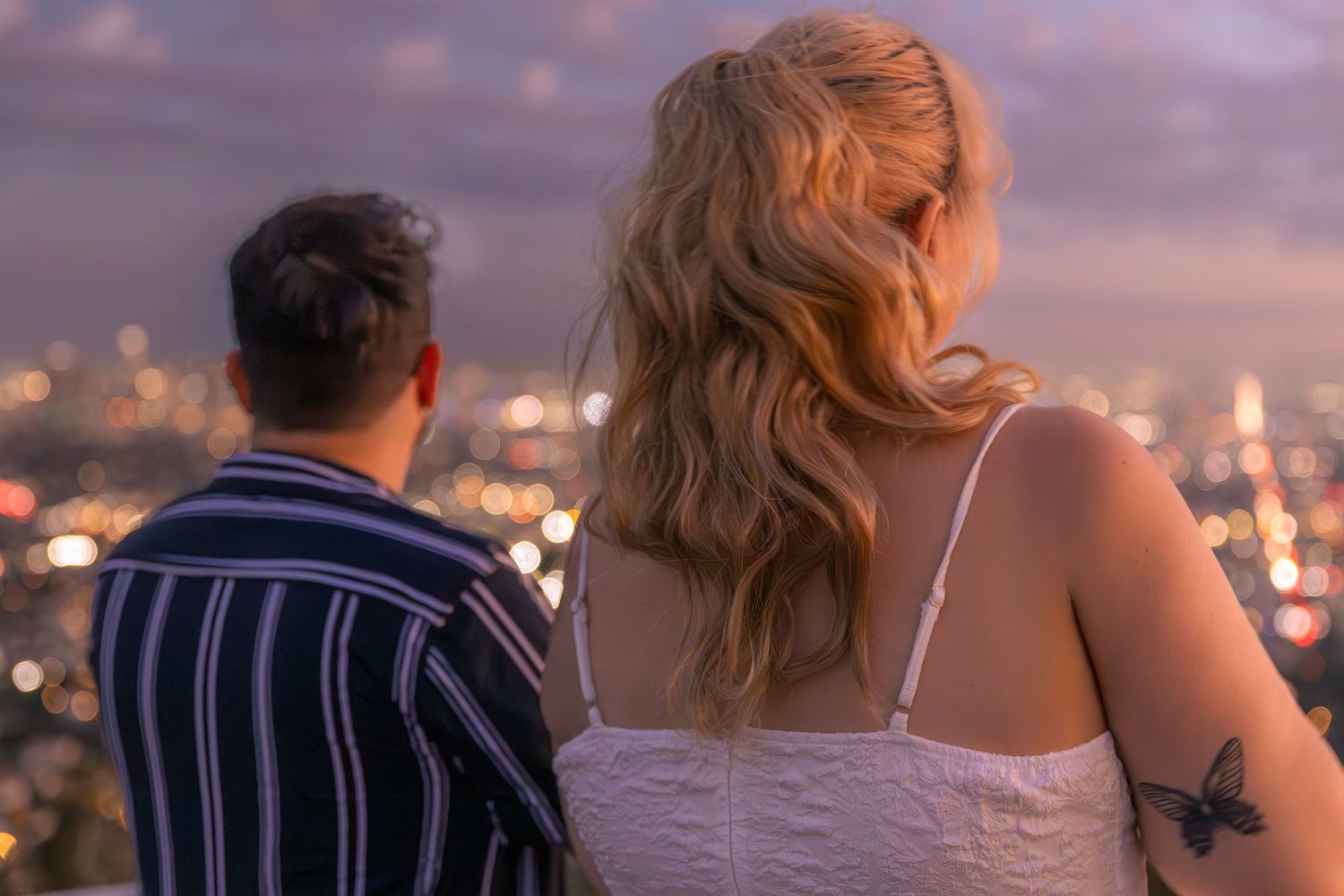 Couple enjoying a romantic sunset moment at a coastal viewpoint with city lights twinkling in the background.