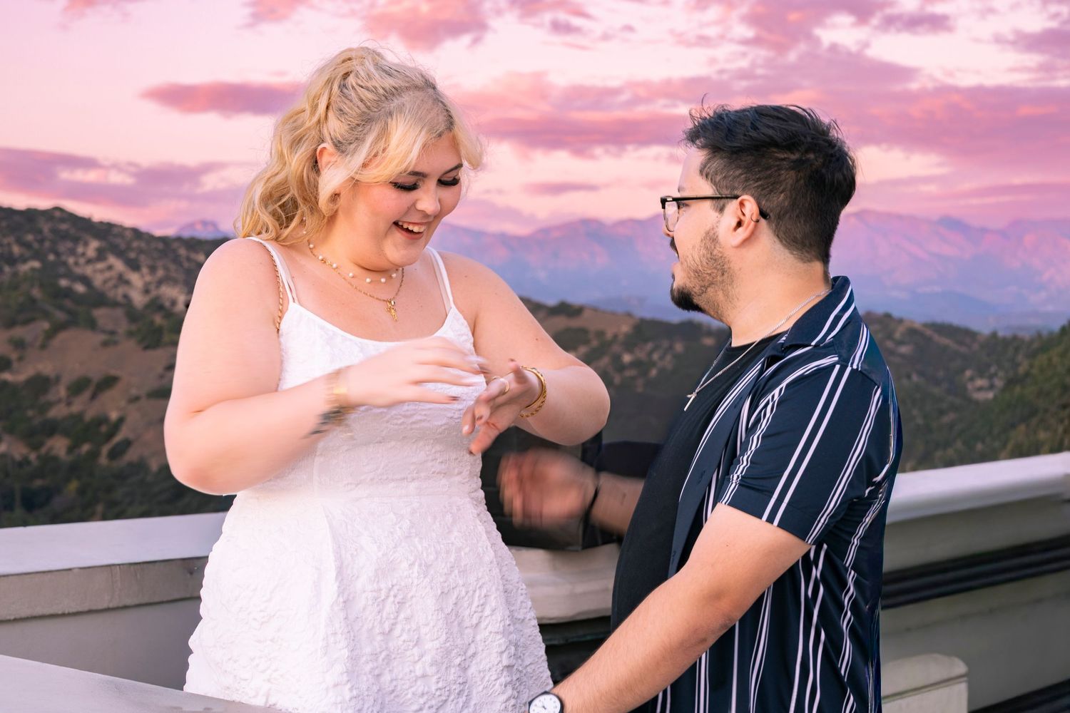 A couple shares a romantic moment on a balcony during a pink and purple sunset in the mountains.