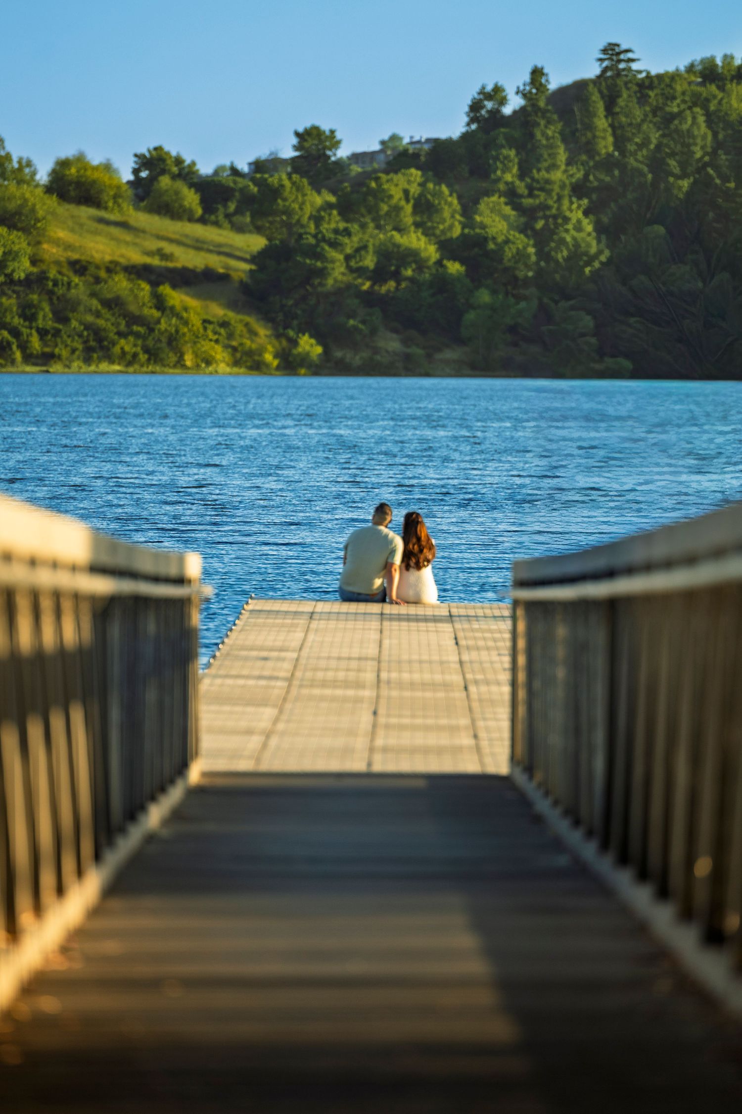 A wooden dock leads to a serene lake view where two people sit together at the end in golden sunlight.