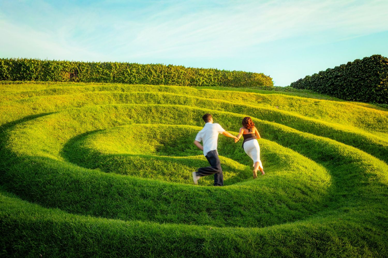Couple running hand-in-hand up spiral grass terraces in a scenic green landscape.