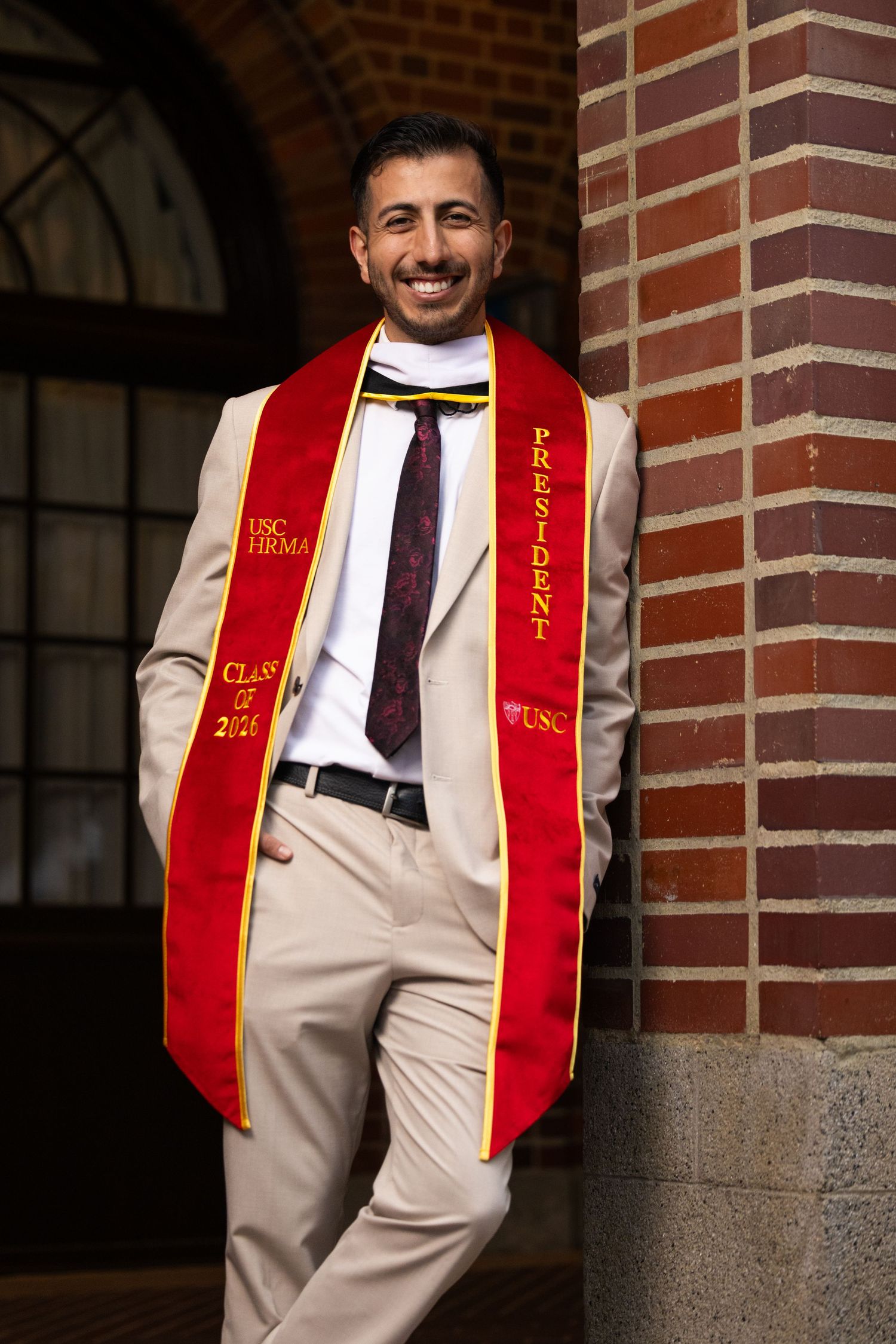 USC graduate smiling in tan suit with red and gold PRESIDENT stole, leaning against brick column.