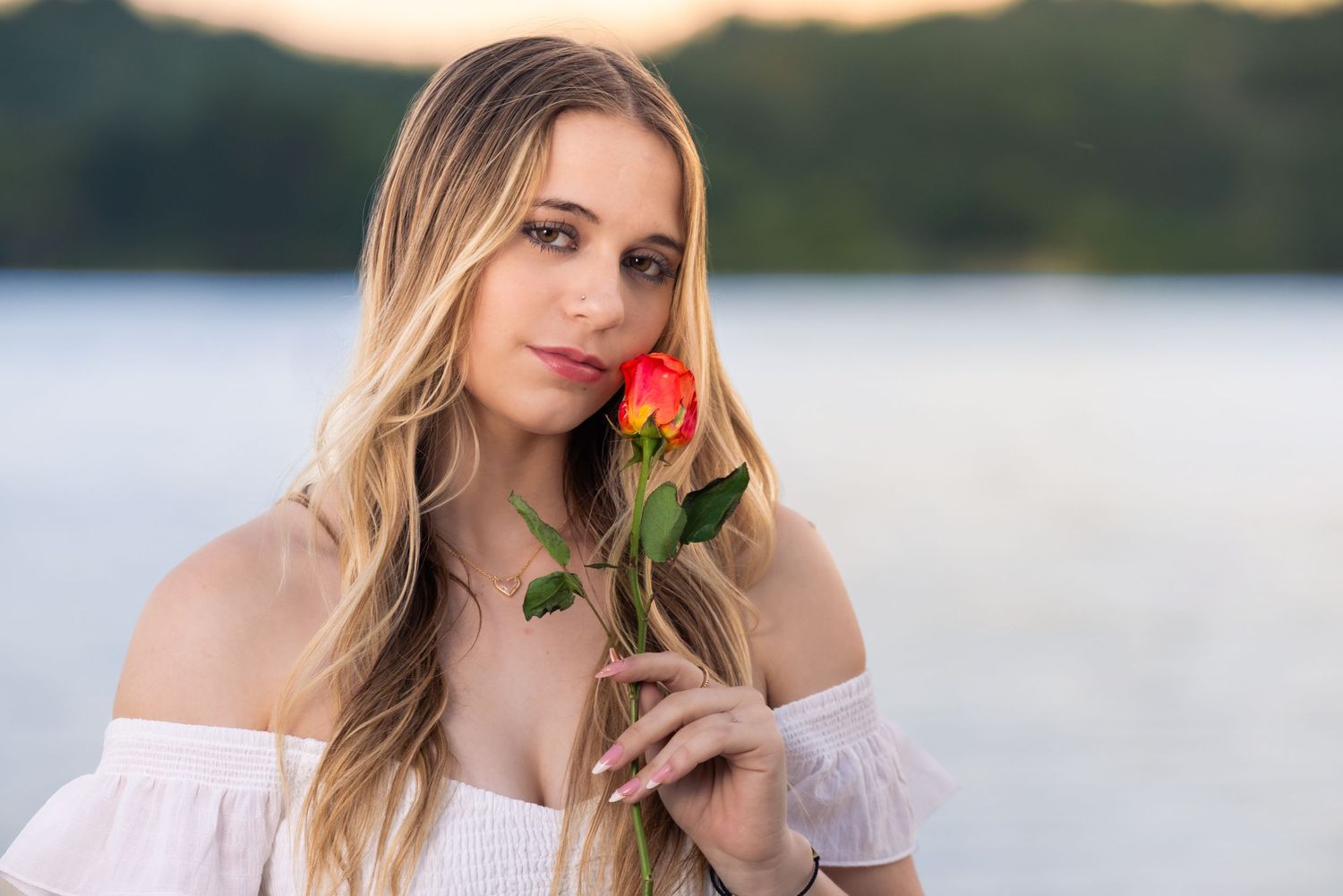 Young blonde woman in white off-shoulder top holds a red rose near her face by a serene lake at sunset.