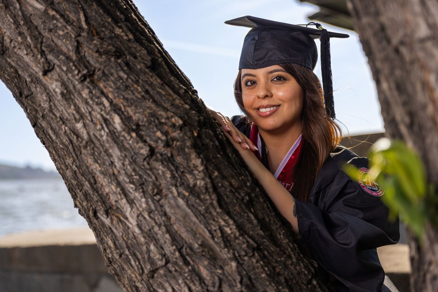 Young woman in graduation cap and gown smiling beside a tree, celebrating her academic achievement outdoors.