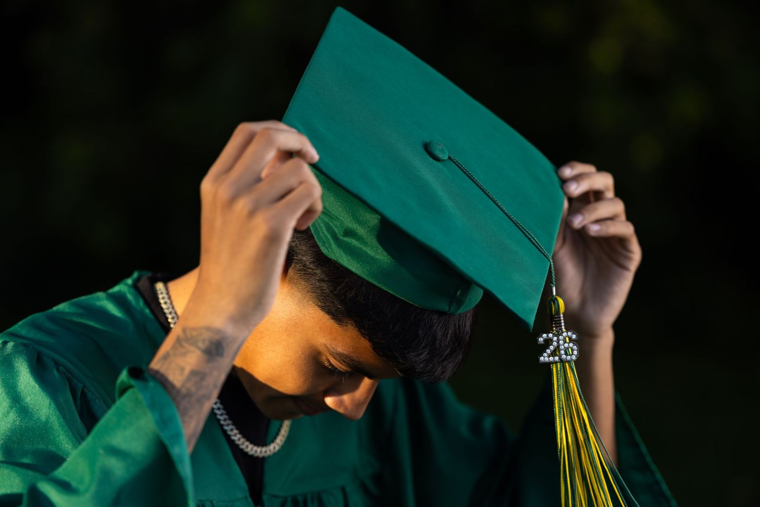 Graduate in green cap and gown adjusting mortarboard tassel, class of 26 graduation ceremony outdoors.