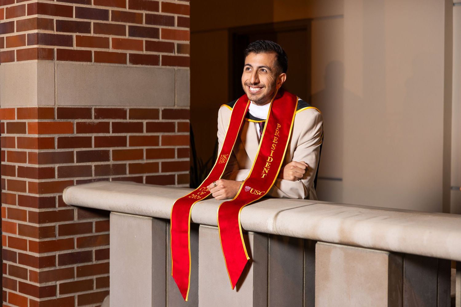 Graduate in white suit with red sash leans on railing, smiling, beside brick columns during graduation celebration.