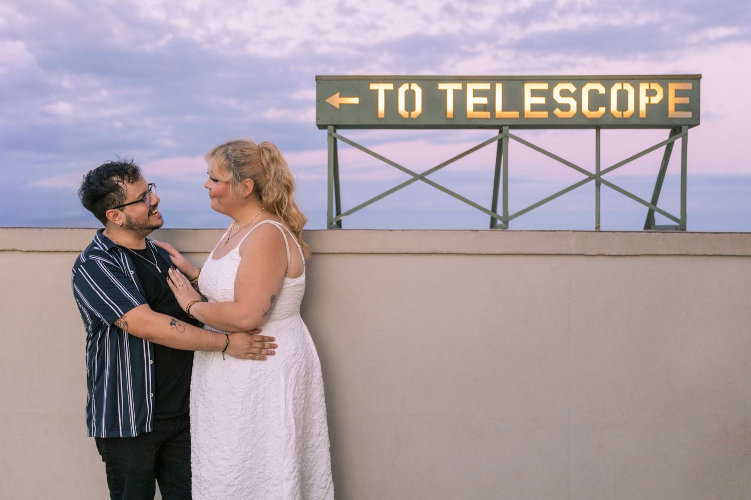 A couple shares a romantic moment at sunset on a scenic overlook with city views in the background.
