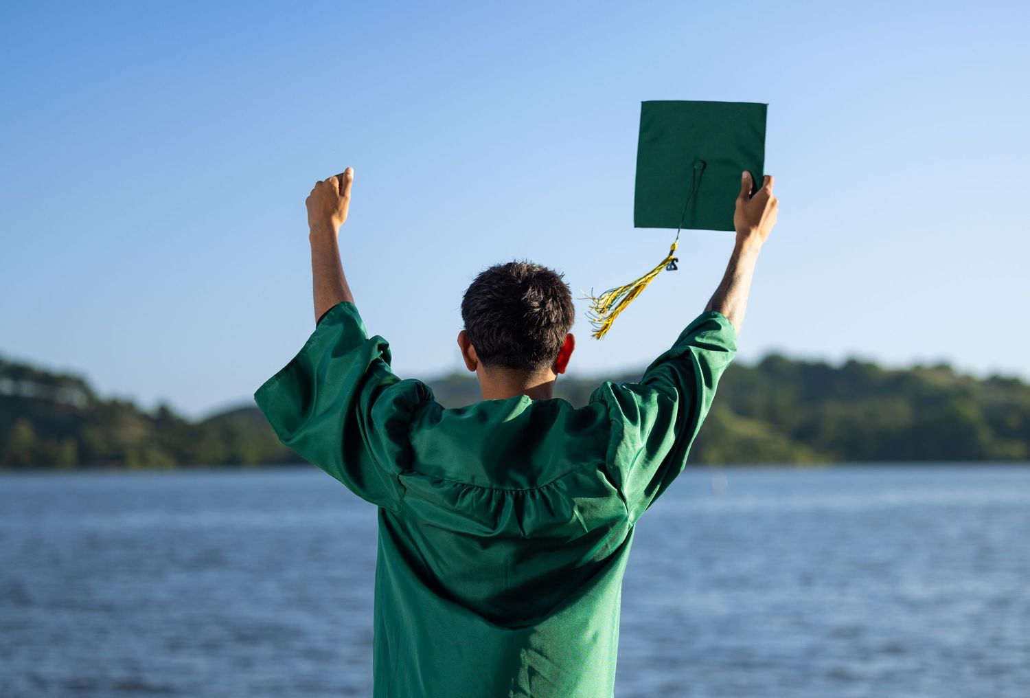 Graduate in green gown raises cap triumphantly by a lake under clear blue sky.