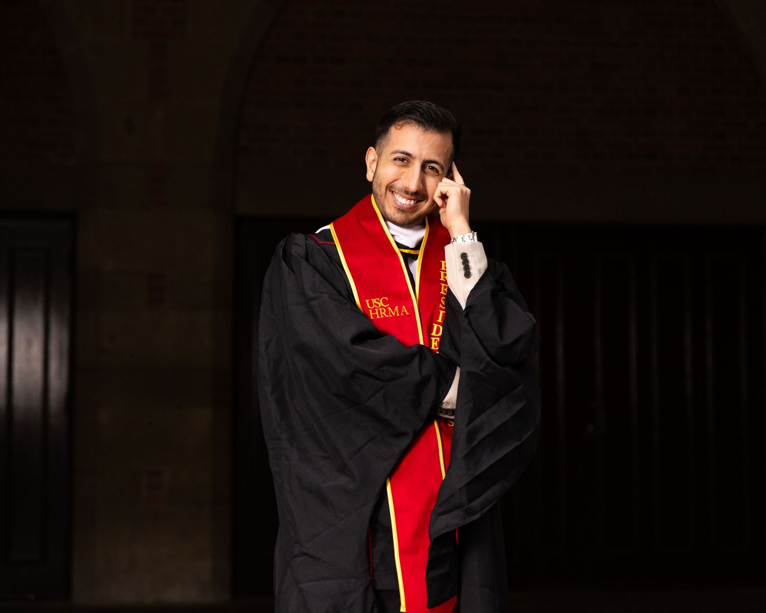 Smiling graduate in black cap and gown with red and gold stole poses under archway.