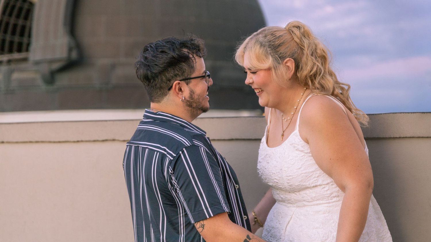 Couple in formal attire shares tender moment on outdoor terrace at sunset.