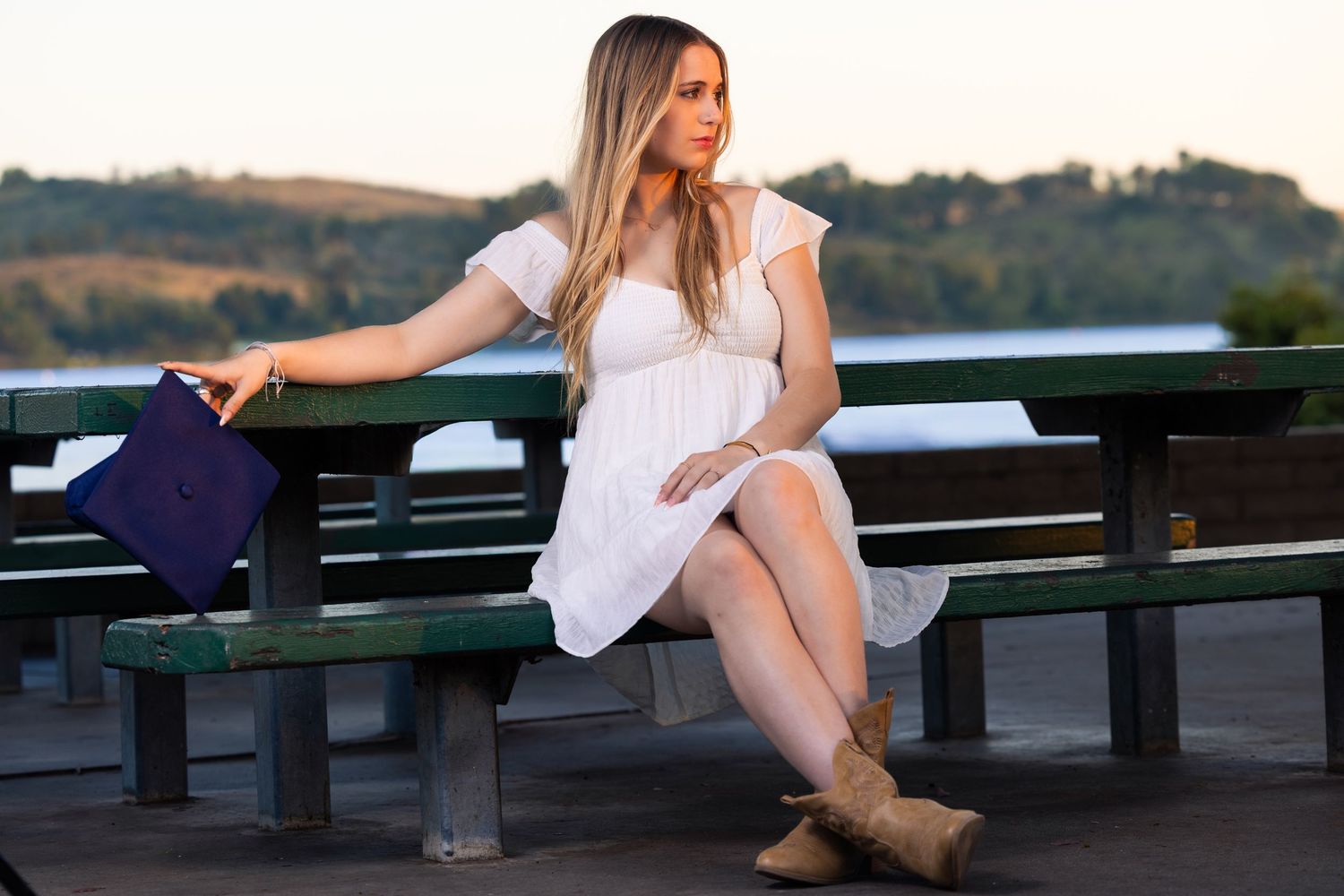 Young woman in white dress and boots sits on park bench holding purple graduation cap at sunset.